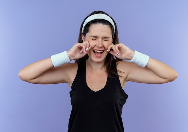 Young fitness woman in headband closing ears happy and excited standing over blue background