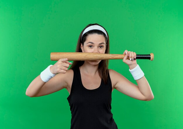 young fitness woman in headband holding baseball bat  pointing with finger  smiling standing over green wall