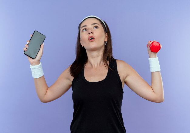 young fitness woman in headband holding smartphone and dumbbell looking confused standing over blue wall