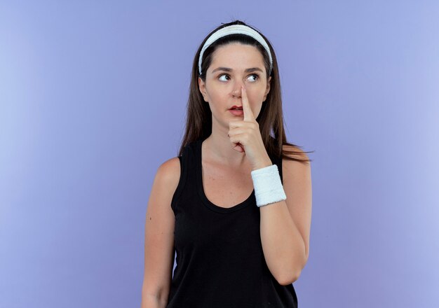 young fitness woman in headband looking aside pointing with finger to her nose standing over blue wall