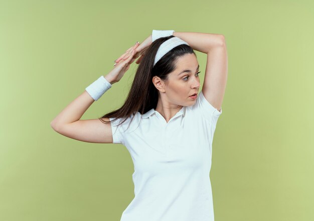 Young fitness woman in headband looking aside stretching her hands standing over light background