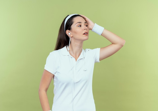 Young fitness woman in headband looking aside with confident expression standing over light wall