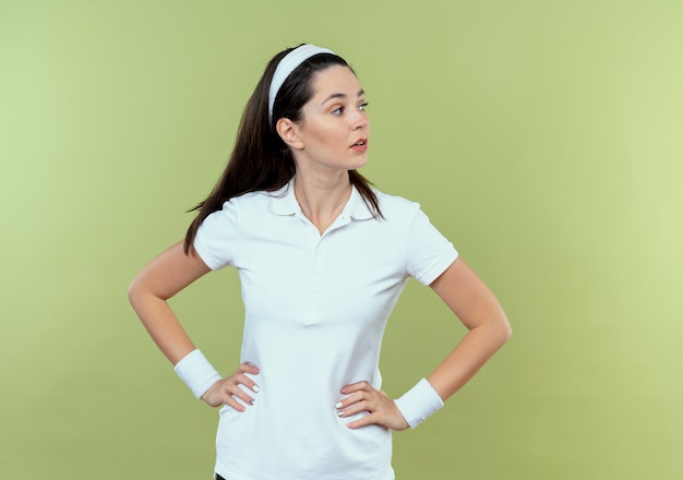 young fitness woman in headband looking aside with confident expression with arms at hip standing over light wall