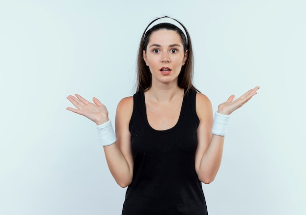 Free photo young fitness woman in headband looking at camera confused and uncertain spreading arms to the side standing over white background