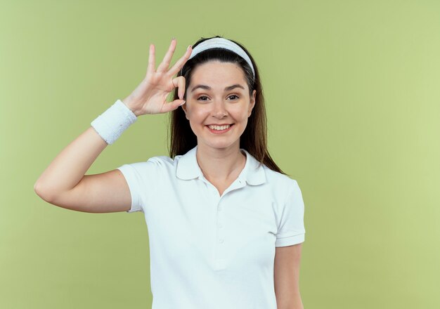 Young fitness woman in headband looking at camera smiling cheerfully showing ok sign standing over light background