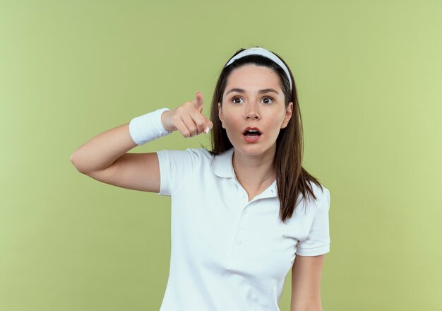 Young fitness woman in headband looking surprised pointing with finger at something standing over light background