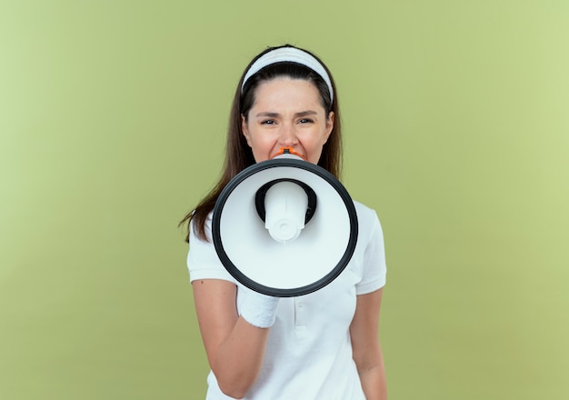 young fitness woman in headband shouting to megaphone loudly standing over light wall