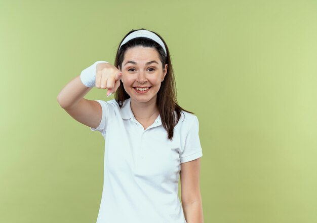 Young fitness woman in headband smiling cheerfully pointing with finger to camera standing over light background