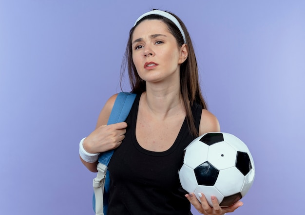 Young fitness woman in headband with backpack holding soccer ball looking confused standing over blue background