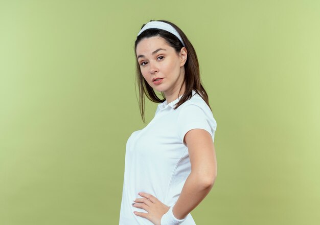 Free photo young fitness woman in headband  with confident expression with arms at hip standing over light wall