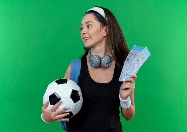 Young fitness woman in headband with headphones and backpack holding soccer ball and air tickets looking aside smiling standing over green background