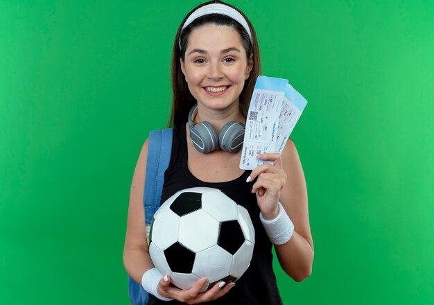 Young fitness woman in headband with headphones and backpack holding soccer ball and air tickets looking at camera smiling with happy face standing over green background