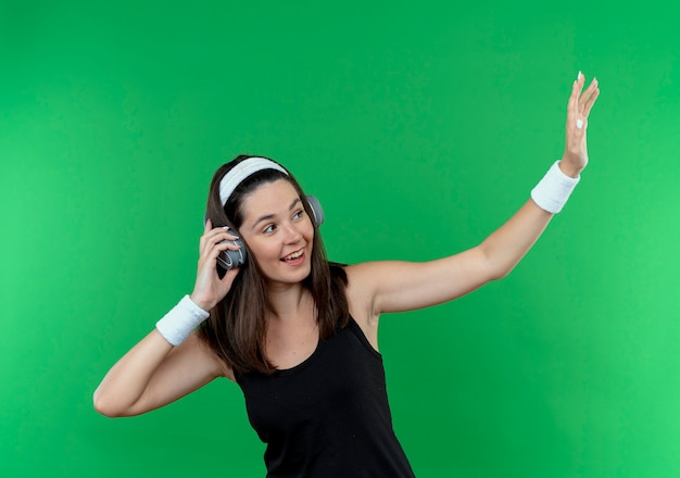 young fitness woman in headband with headphones looking aside smiling cheerfully waving with a hand standing over green wall