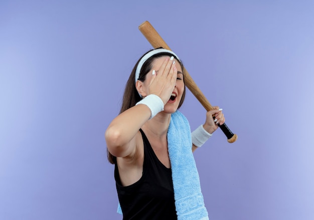 Young fitness woman in headband with towel on her shoulder holding baseball bat covering one eye with a hand smiling standing over blue background