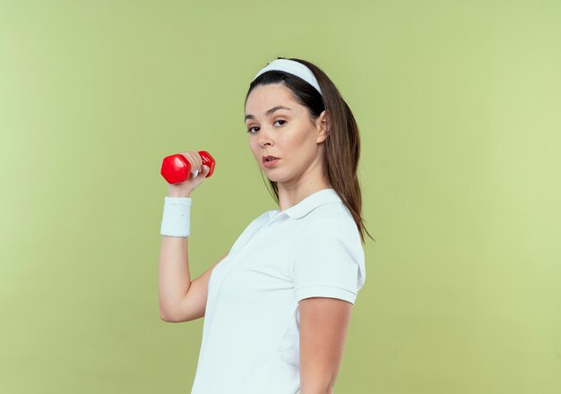 Young fitness woman in headband working out with dumbbell looking at camera with serious face standing over light background