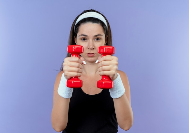 Free Photo young fitness woman in headband working out with dumbbells looking confident standing over blue background