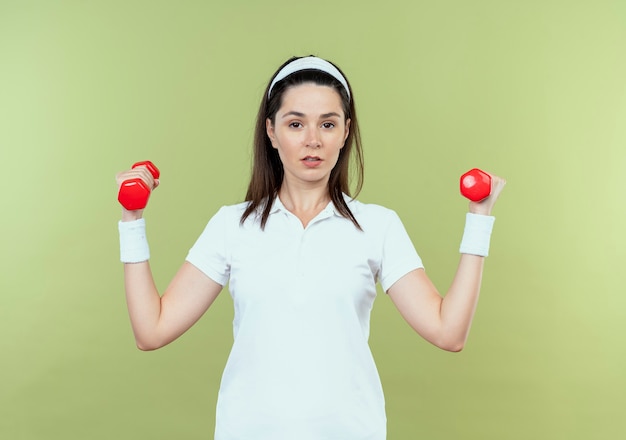 Young fitness woman in headband working out with dumbbells looking confident standing over light wall