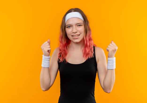 Young fitness woman in sportswear clenching fists happy and exited rejoicing her success standing over orange wall