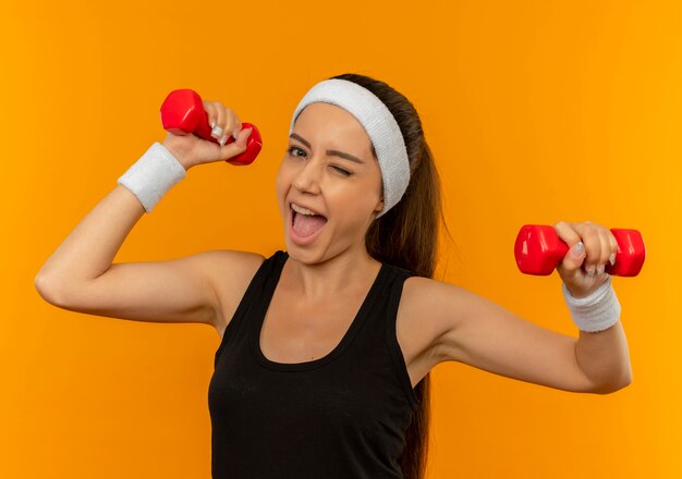 Young fitness woman in sportswear doing exercises using two dumbbells smiling cheerfully standing over orange wall