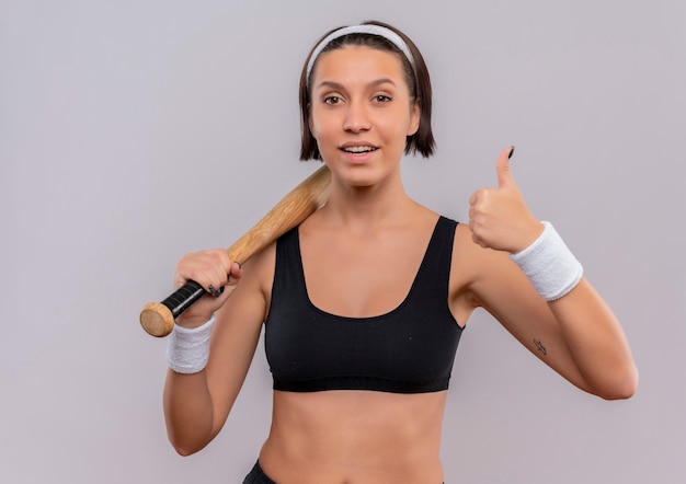 Young fitness woman in sportswear holding baseball bat smiling confident showing thumbs up standing over white wall