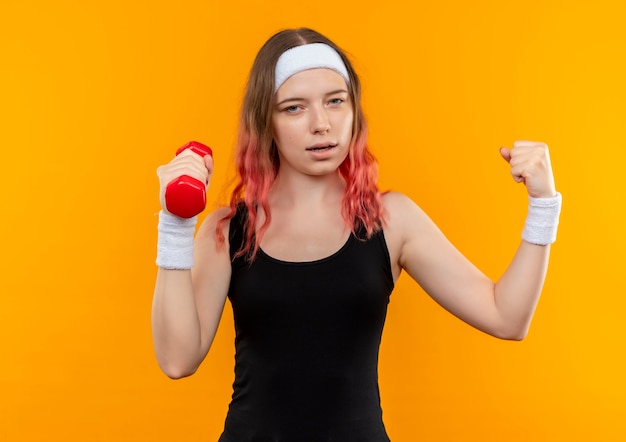 Young fitness woman in sportswear holding dumbbell raising fist like a winner standing over orange wall