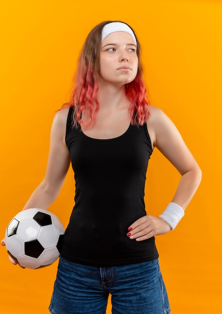 Free Photo young fitness woman in sportswear holding soccer ball looking aside with serious face standing over orange wall