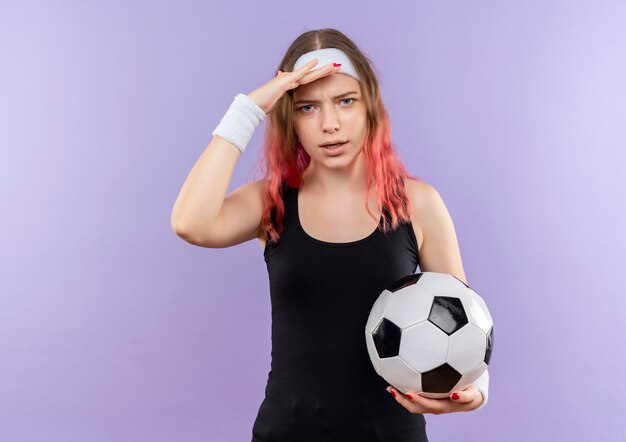 Young fitness woman in sportswear holding soccer ball with hand over head standing over purple wall