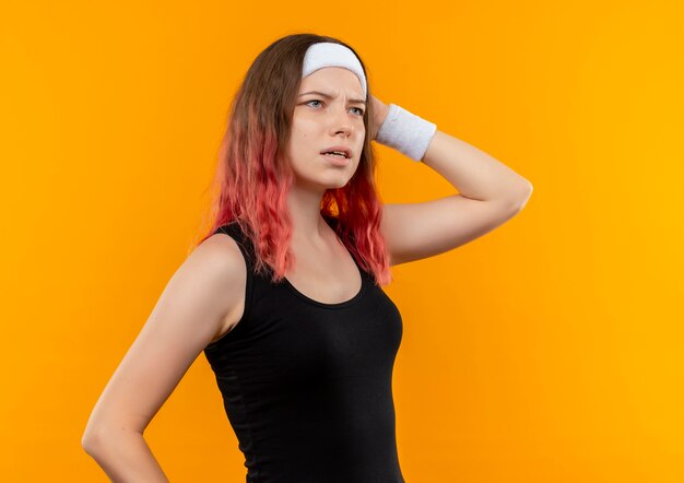 Young fitness woman in sportswear looking aside confused and very anxious standing over orange wall