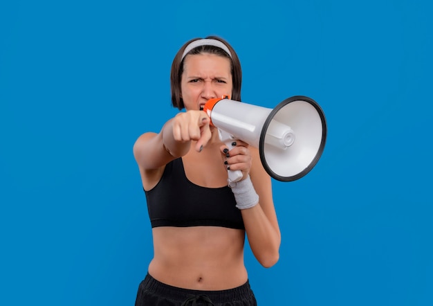 Young fitness woman in sportswear shouting to megaphone with aggressive expression pointing with index finger to camera standing over blue wall