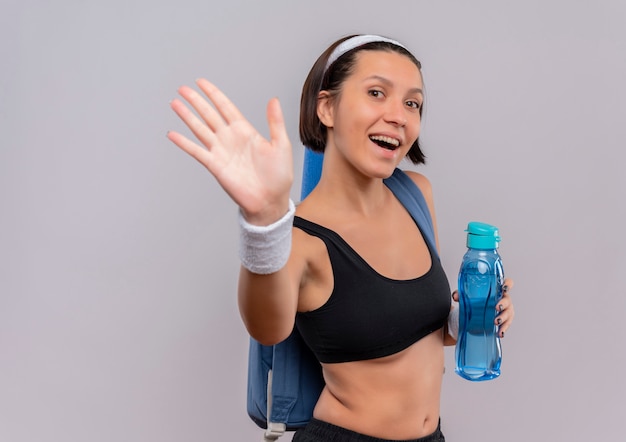 Young fitness woman in sportswear with backpack and yoga mat holding bottle of water smiling waving with hand standing over white wall