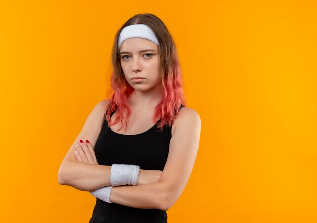 Young fitness woman in sportswear with crossed arms on chest with serious face standing over orange wall