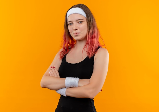 Young fitness woman in sportswear with crossed arms with confident smile on face standing over orange wall