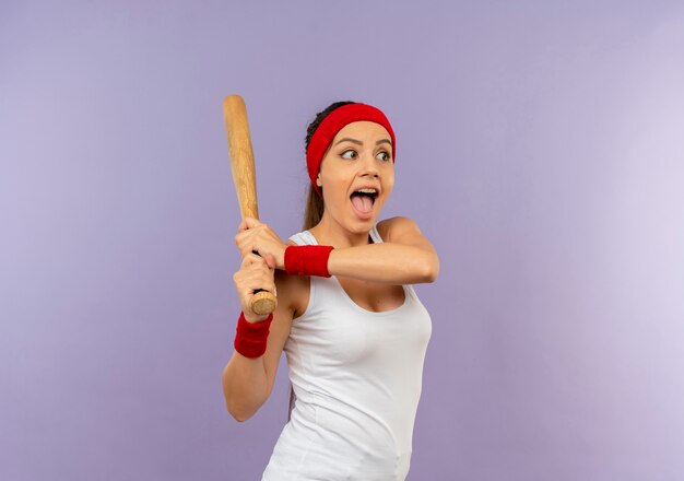 Young fitness woman in sportswear with headband holding baseball bat playing happy and excited standing over grey wall