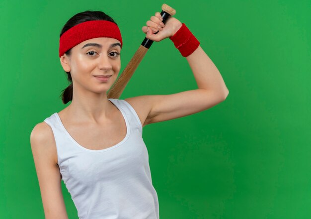 Young fitness woman in sportswear with headband holding baseball bat with confident smile standing over green wall