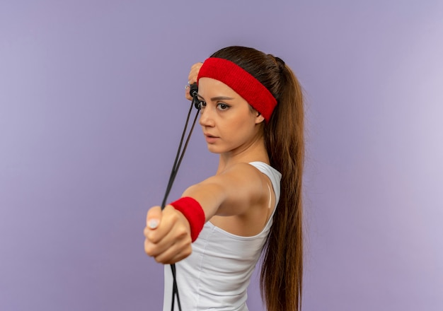 Free photo young fitness woman in sportswear with headband holding skipping rope like aiming with a bow and arrow standing over grey wall