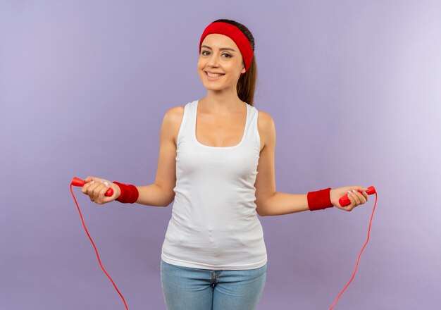 Young fitness woman in sportswear with headband holding skipping rope smiling cheerfully going to jump standing over grey wall