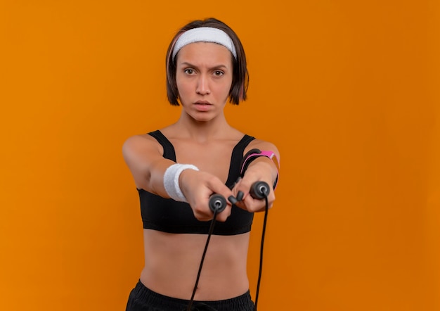 Young fitness woman in sportswear with headband holding skipping rope with confident expression standing over orange wall