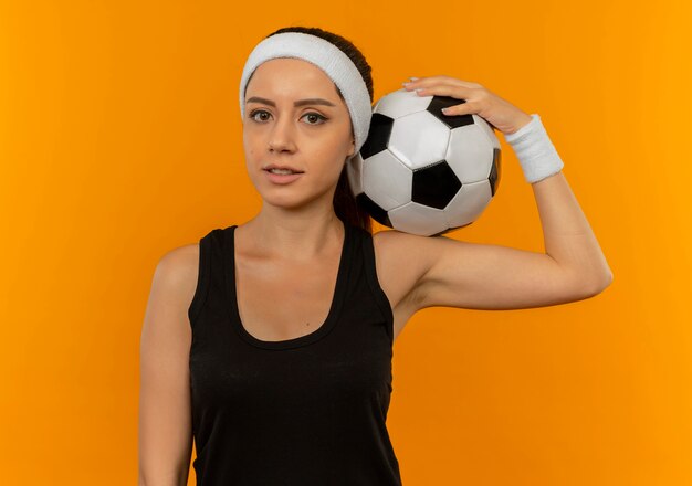 Young fitness woman in sportswear with headband holding soccer ball looking confident standing over orange wall