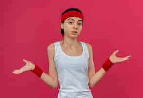 Free photo young fitness woman in sportswear with headband looking confused and uncertain spreading arms to the sides having no answer standing over pink wall