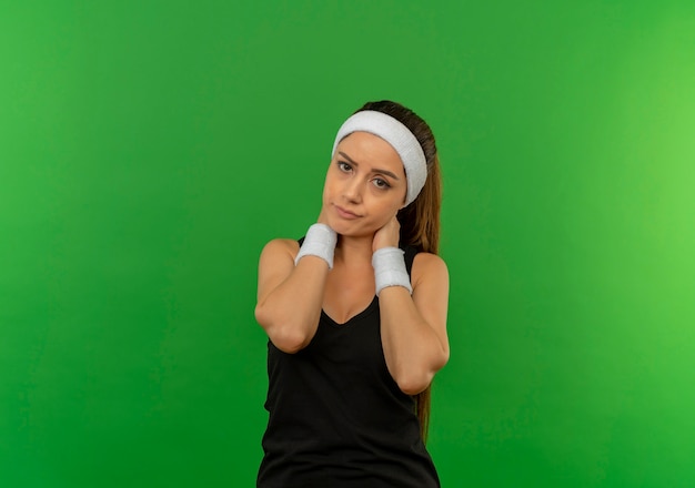 Young fitness woman in sportswear with headband looking unwell touching her neck having pain standing over green wall