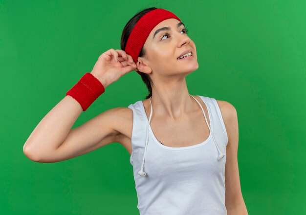 Young fitness woman in sportswear with headband looking up smiling feeling positive emotions touching her headband standing over green wall