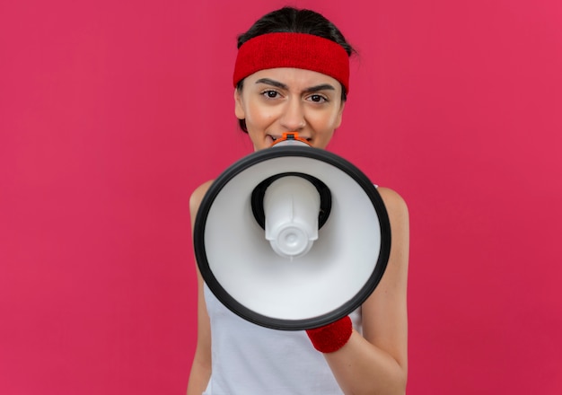 Free photo young fitness woman in sportswear with headband shouting to megaphone with angry face standing over pink wall