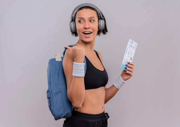 Young fitness woman in sportswear with headphones on head with backpack holding air ticket looking aside pointing back with thumb standing over white wall