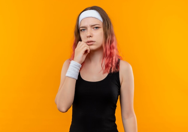 Young fitness woman in sportswear with pensive expression on face, thinking standing over orange wall