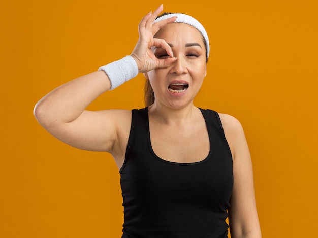 Young fitness woman with headband and armbands doing ok sign looking through fingers smiling  