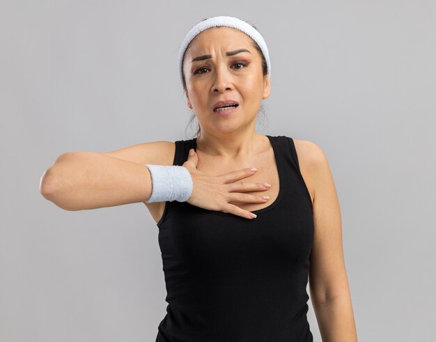 Young fitness woman with headband and armbands  feeling unwell holding hand on her chest standing over white wall