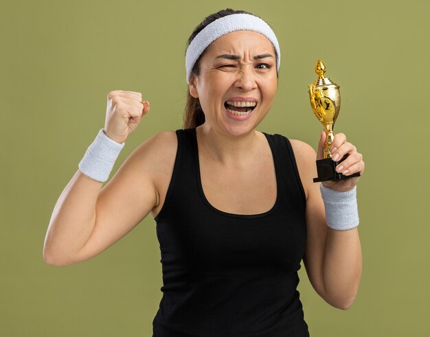 Young fitness woman with headband and armbands holding trophy angry and frustrated clenching fist  