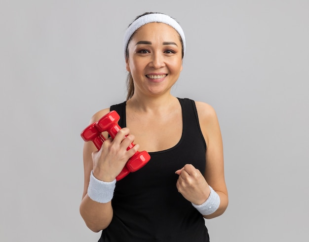 Young fitness woman with headband holding dumbbells doing exercises clenching fist happy and excited  standing over white wall