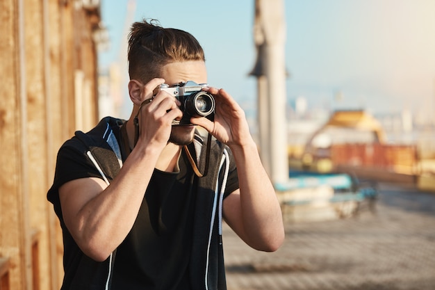 Free photo young focused european guy standing in harbour looking through camera while taking pictures of sea or yachts, walking along city to gather cool photos for magazine. talented cameraman searching angle