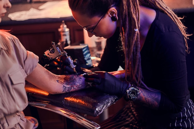 Free photo young focused tattoo master is makining tattoo on client's hand at dark tattoo salon.